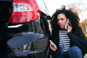 Angry young woman standing by vandalised car in car park reporting damage to insurance company using mobile phone
