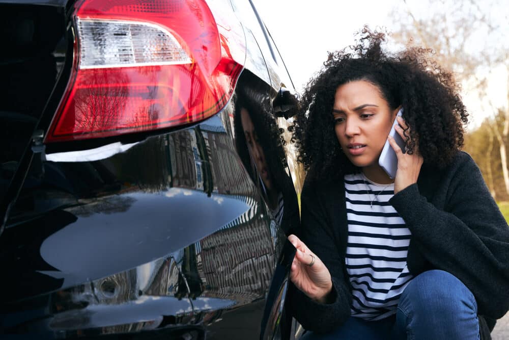 Angry young woman standing by vandalised car in car park reporting damage to insurance company using mobile phone