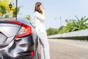 Upset unhappy Asian woman have a car accident or crashed during her road trip, woman driver sitting beside the crashed or broken car and using cell phone calling to road side assistant service.