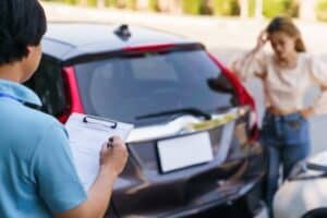 Insurance agent filling out vehicle accident claim form on clipboard, car surveyor documenting vehicle damage while the driver feels upset in background.