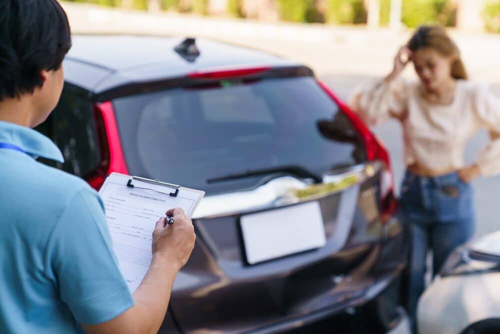 Insurance agent filling out vehicle accident claim form on clipboard, car surveyor documenting vehicle damage while the driver feels upset in background.