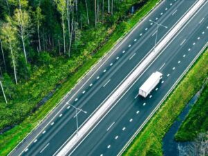 Aerial drone view of freight transport semi-trucks and cars driving on highway asphalt road through green summer woods.