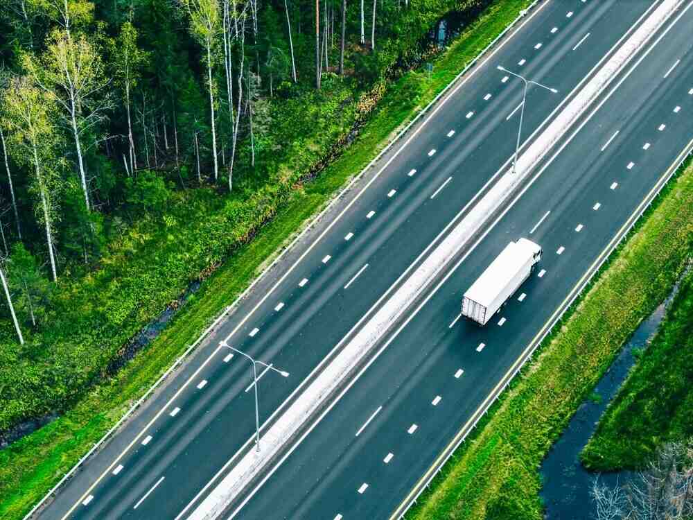 Aerial drone view of freight transport semi-trucks and cars driving on highway asphalt road through green summer woods.